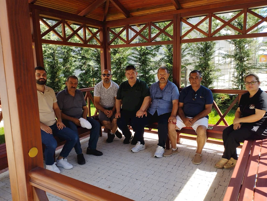Ahşap kamelyada oturan 7 kişilik Bahçekentli grup, dostça ve samimi bir havada objektife poz veriyor. Hepsi Bahçekent’in köklü isimleri. Arka planda yemyeşil çam ağaçları ve tertemiz çevre düzenlemesi görülüyor. Fotoğraf, Başakşehir Bizim Çınarlar Yaşam Merkezi'nde çekildi. Ortam huzur, güven ve dostluğun birleştiği bir tabloyu yansıtıyor.
