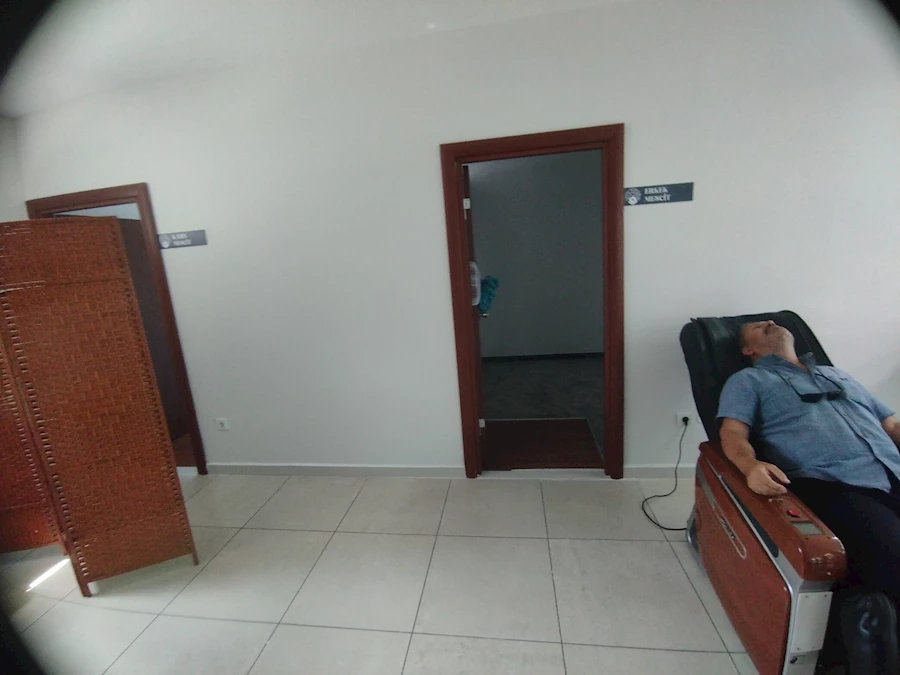 Fotoğrafta bir emekli vatandaş, Başakşehir Bizim Çınarlar Yaşam Merkezi’nde bulunan konforlu masaj koltuğunda gözleri kapalı şekilde dinleniyor. Arka planda “Erkek Mescit” ve “Bayan Mescit” yazılı kapılar, sağda ise sade beyaz duvarlar görülüyor. Koltuk elektrikle çalışıyor ve kullanıcıya sırt, bel ve bacak bölgelerinde rahatlatıcı masaj sunuyor. Bu hizmet tamamen ücretsiz sunuluyor.