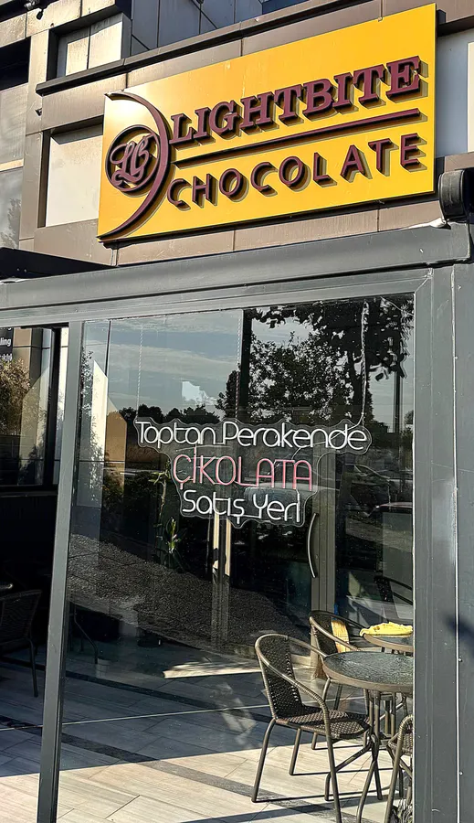 “Bahçekent’te açılan Lightbite Chocolate çikolata mağazasının dış cephesi. Sarı zeminde mor renkte mağaza tabelası ve camda ‘Toptan Perakende Çikolata Satış Yeri’ yazısı görülüyor. Ön tarafta müşteriler için oturma alanı yer alıyor.”
