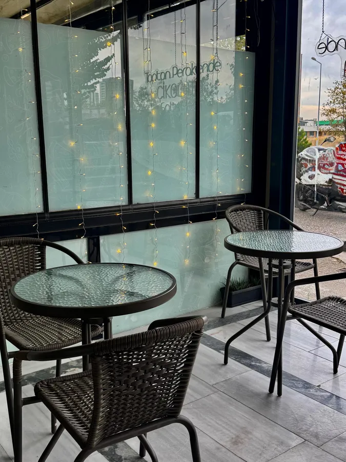 “Bahçekent’teki Lightbite Chocolate mağazasının kafe bölümü. Cam kenarında, ışıklarla süslenmiş alanda yuvarlak cam masalar ve kahverengi sandalyeler bulunuyor. Müşteriler burada kahvelerini içerken çikolataların tadına bakabiliyor.”